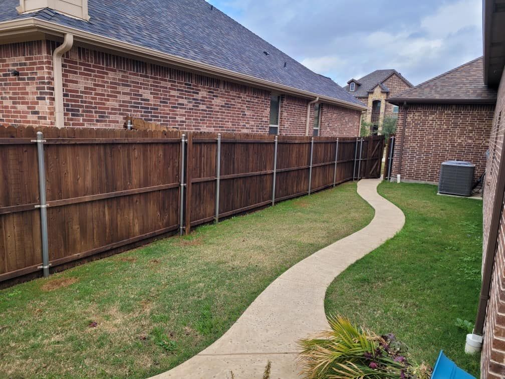 A narrow backyard with a wooden fence, brick walls, and a winding concrete path. Green grass fills the space.