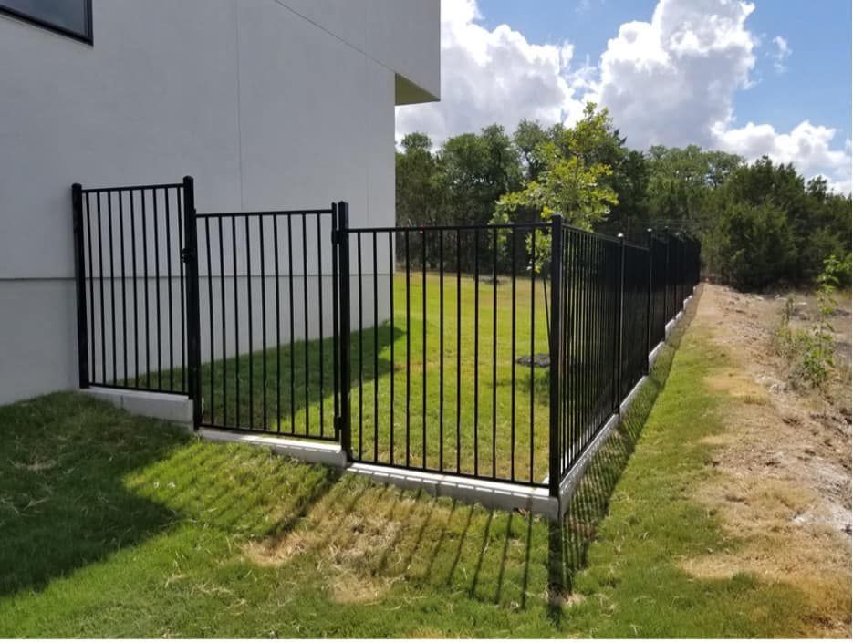 Black metal fence surrounding a small grassy area next to a building, with a gate. Sky and trees are visible in the background.