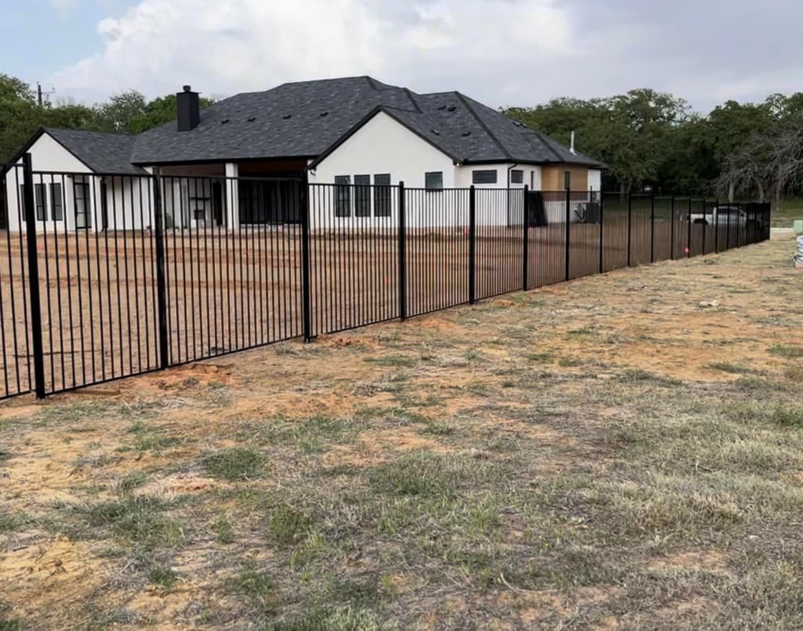 Black metal fence surrounds a beige, grassy yard with a white house in the background. The house has a dark roof.