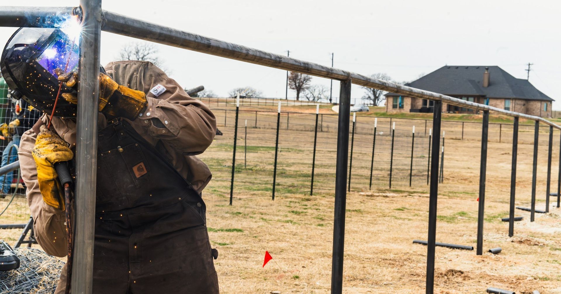 Welder in protective gear attaching railing to a fence post outdoors, near a brown building.