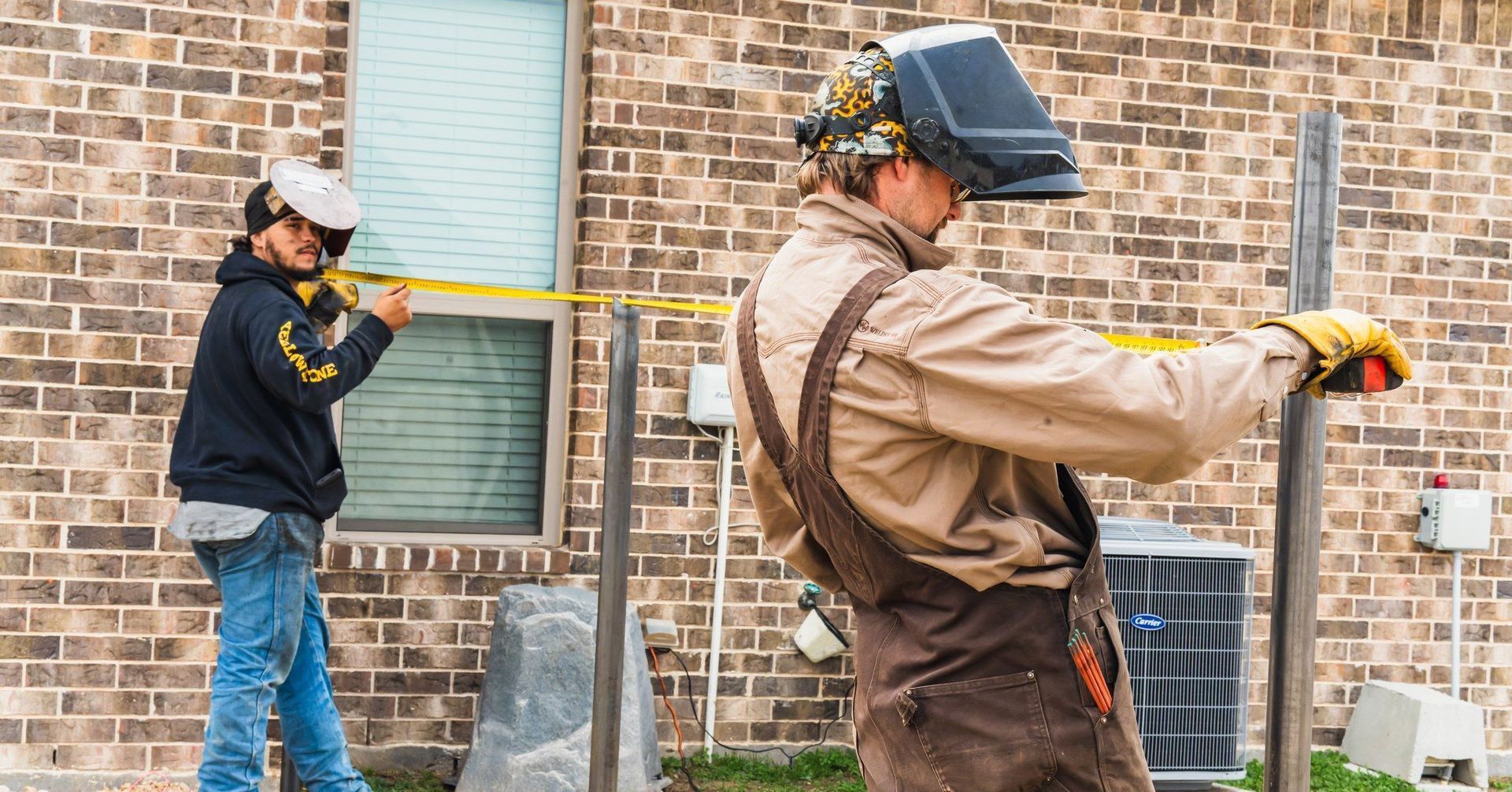 Two workers measuring and welding metal posts near a brick building; one wears a welding mask.