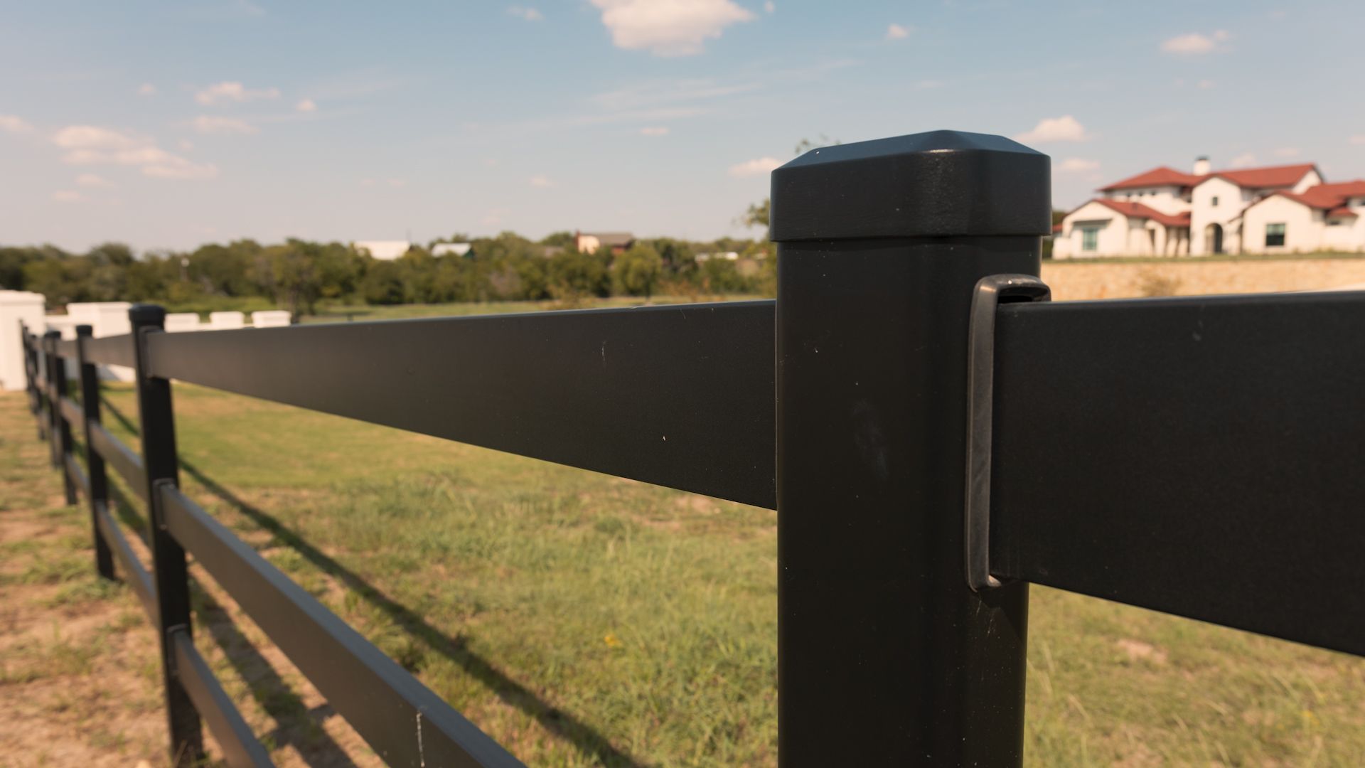 Black fence posts and rails in a grassy field, with a house in the background under a blue sky.