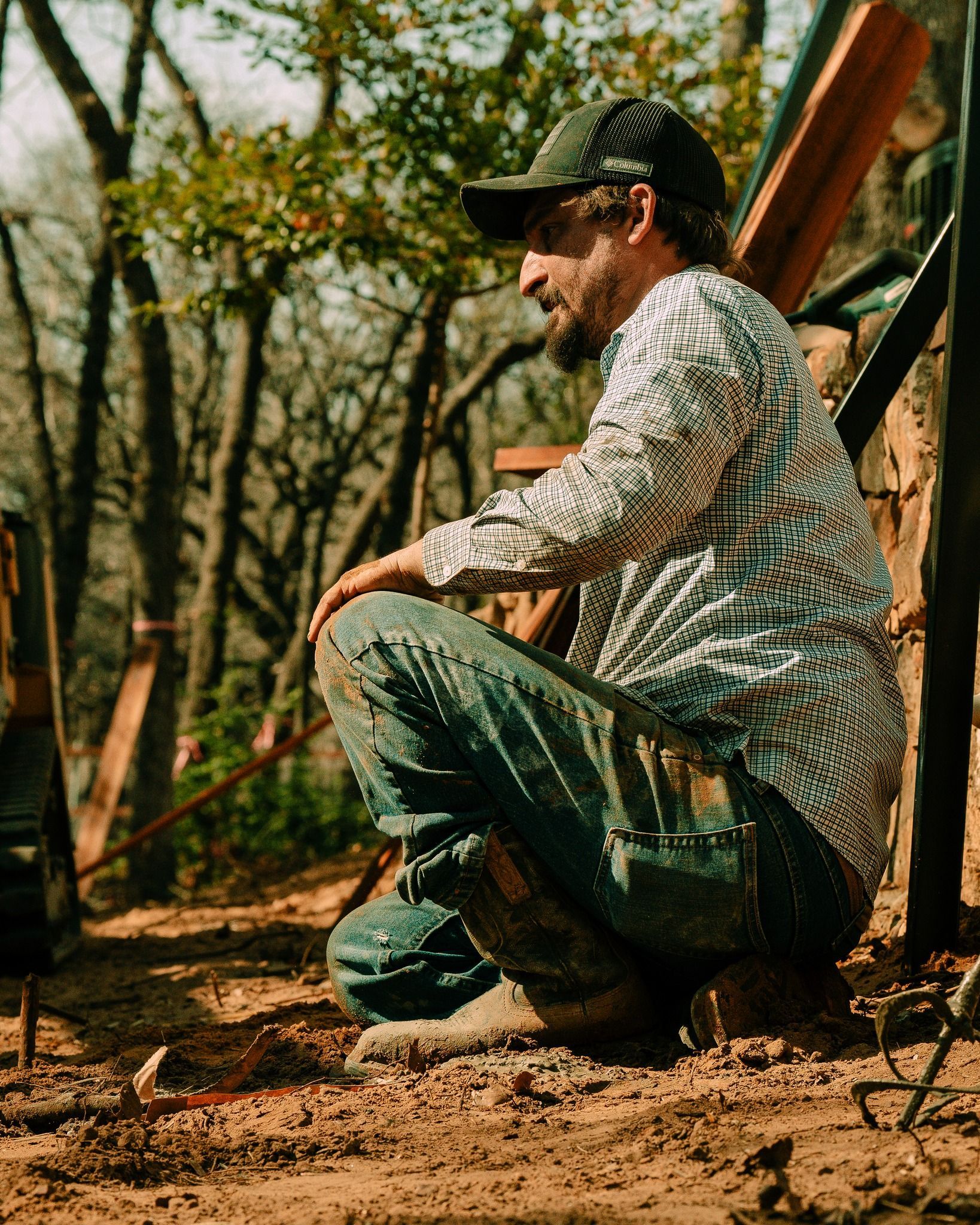Man wearing a hat and patterned shirt, squatting in a wooded area, looking thoughtful. He wears jeans and work boots.