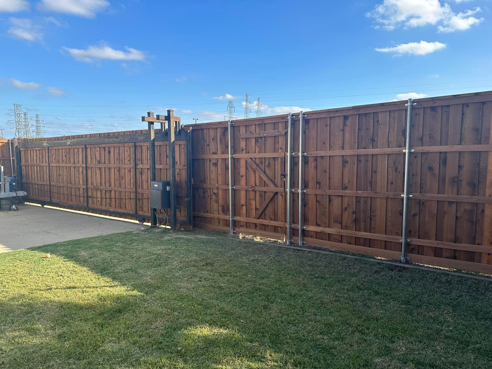 Brown wooden fence with a gate and electrical box in a grassy yard, under a blue sky.