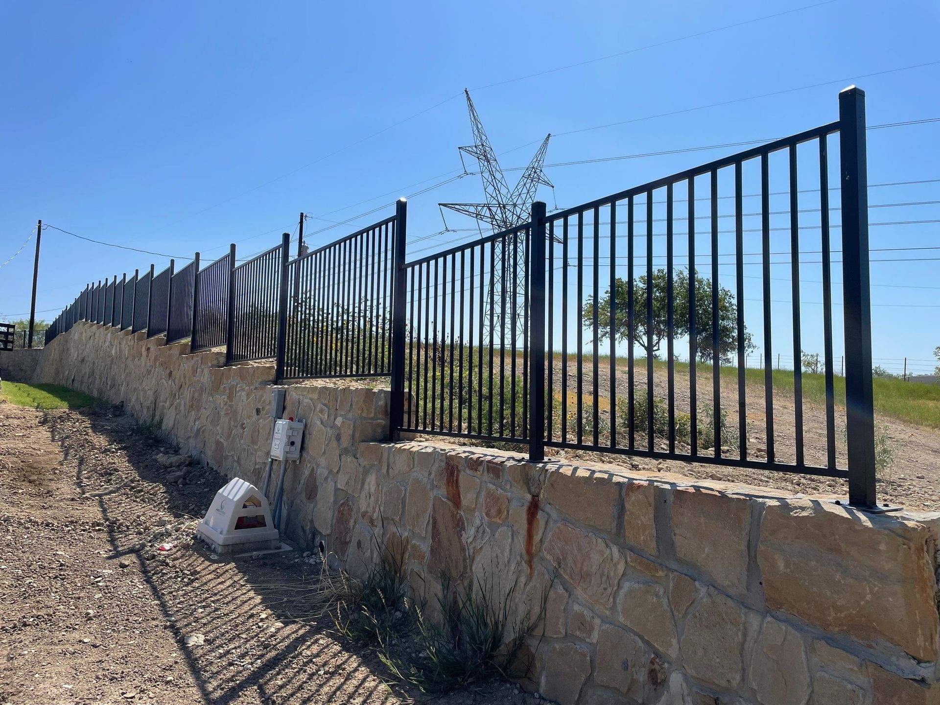Black metal fence atop a stone wall on a sunny day, bordering a grassy area.