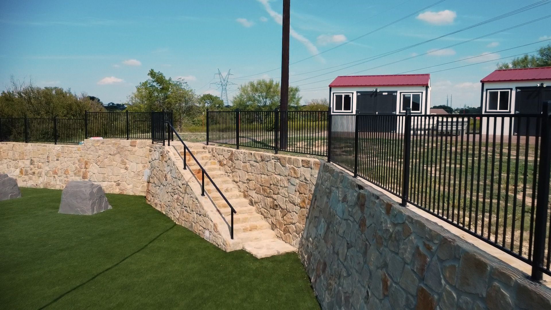 Stone retaining walls with stairs leading up to a black fence and red-roofed buildings under a blue sky.