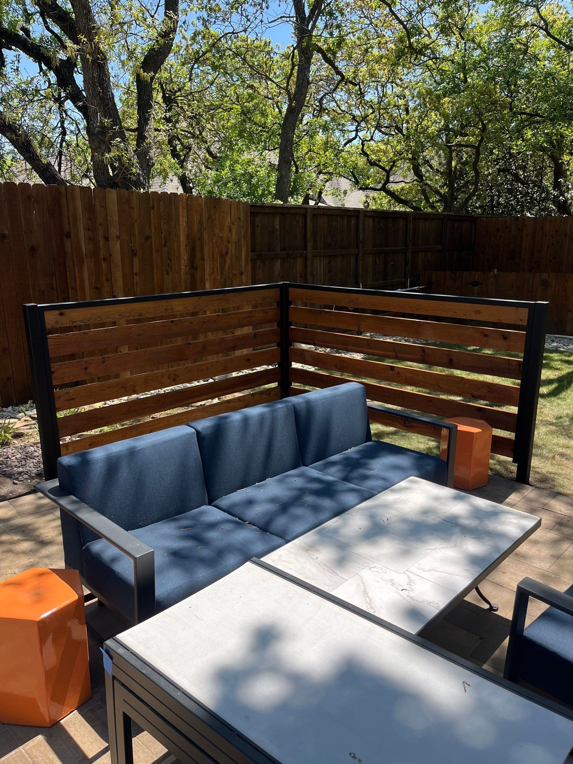 Outdoor patio with a blue sofa, table, and orange stools, set against a wooden privacy fence.