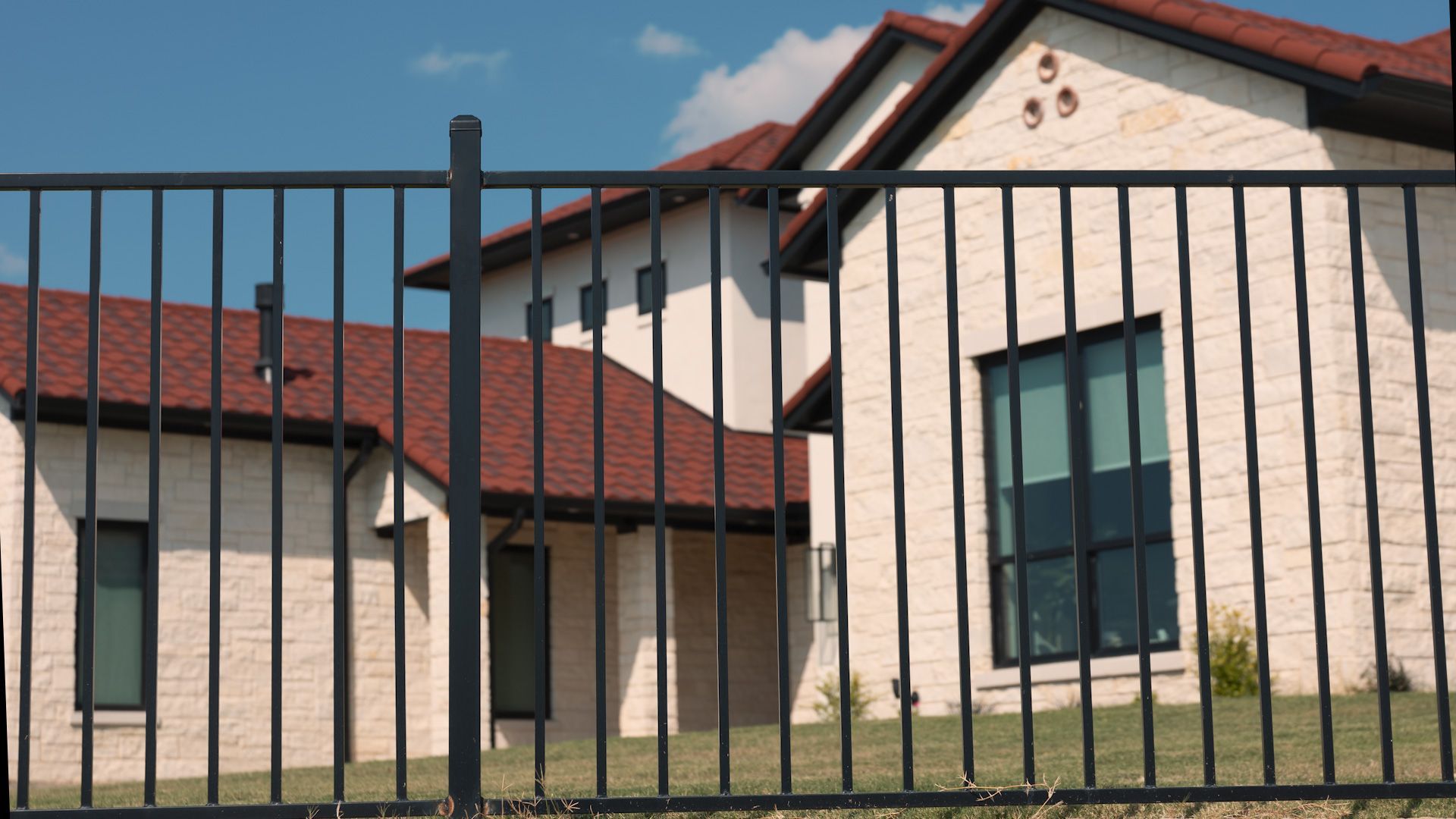 Black metal fence in front of a light brick house with a red roof under a blue sky.