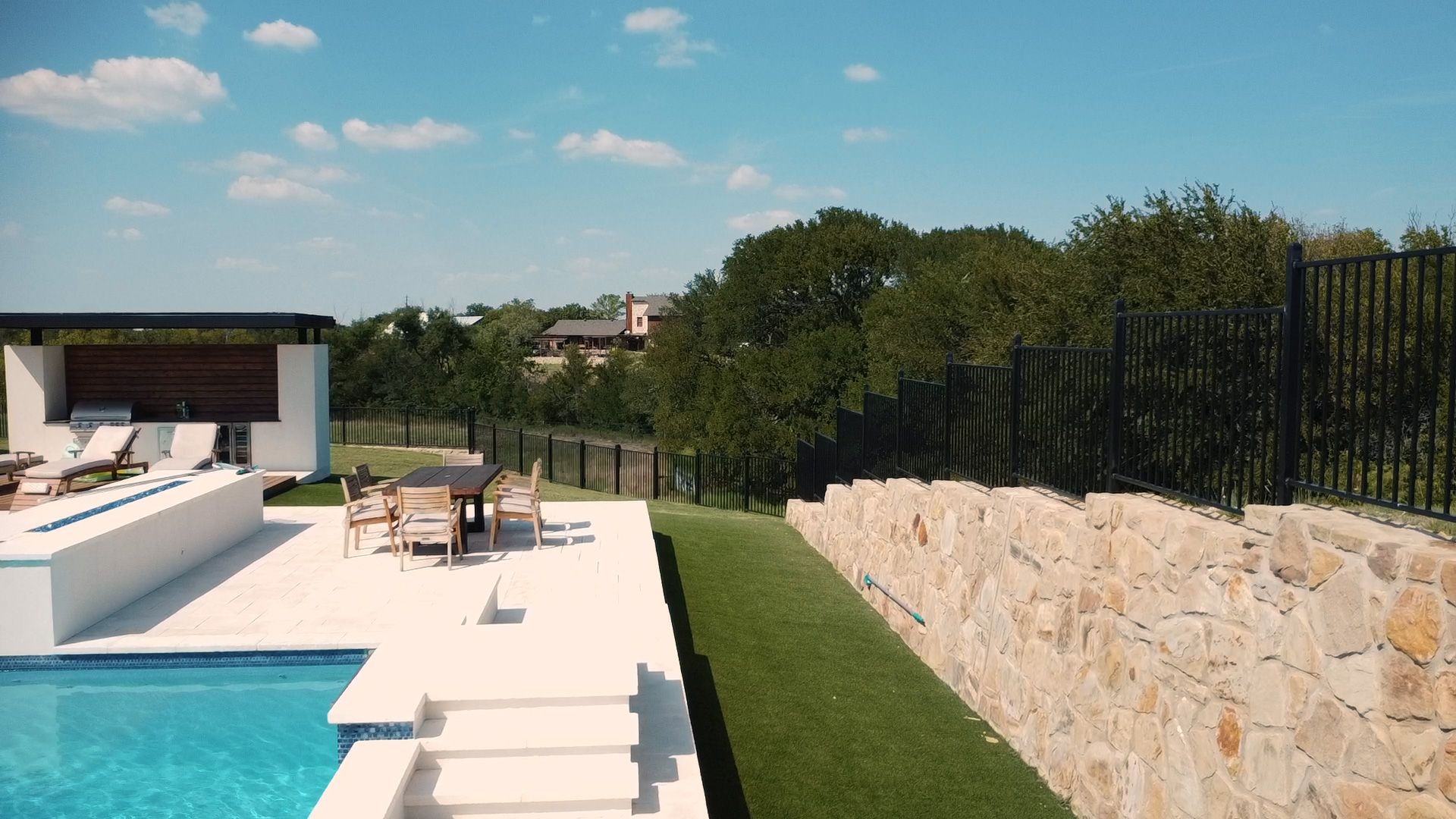 Poolside patio with a pool, table, chairs, and landscaping under a blue sky.