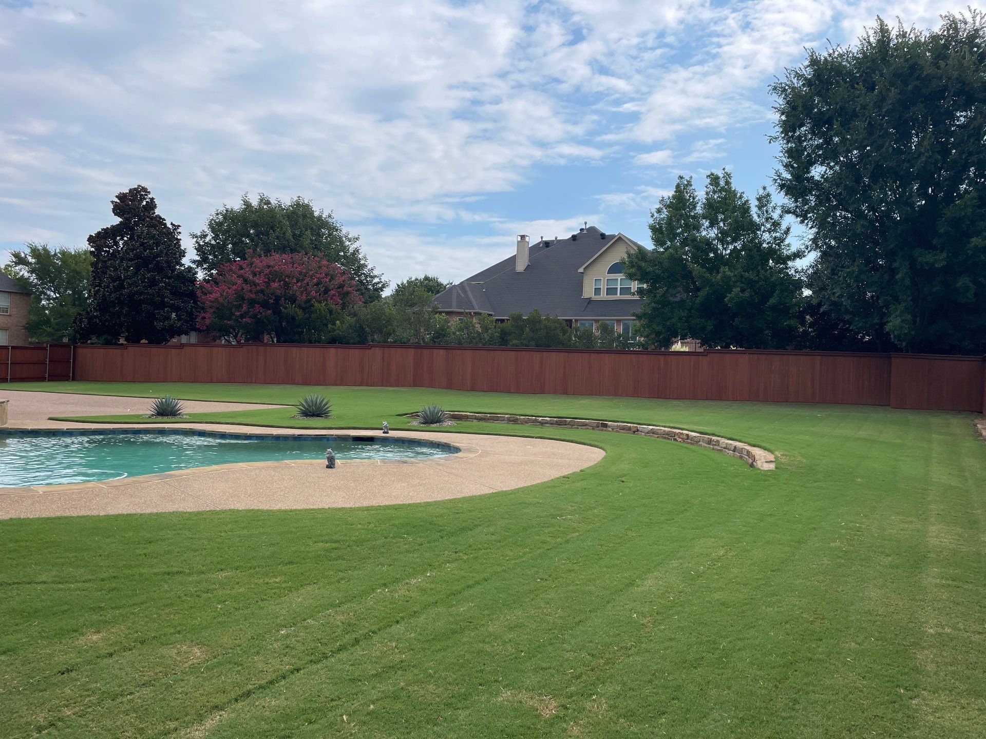 Lush green backyard with a pool, red fence, trees, and a house under a cloudy sky.