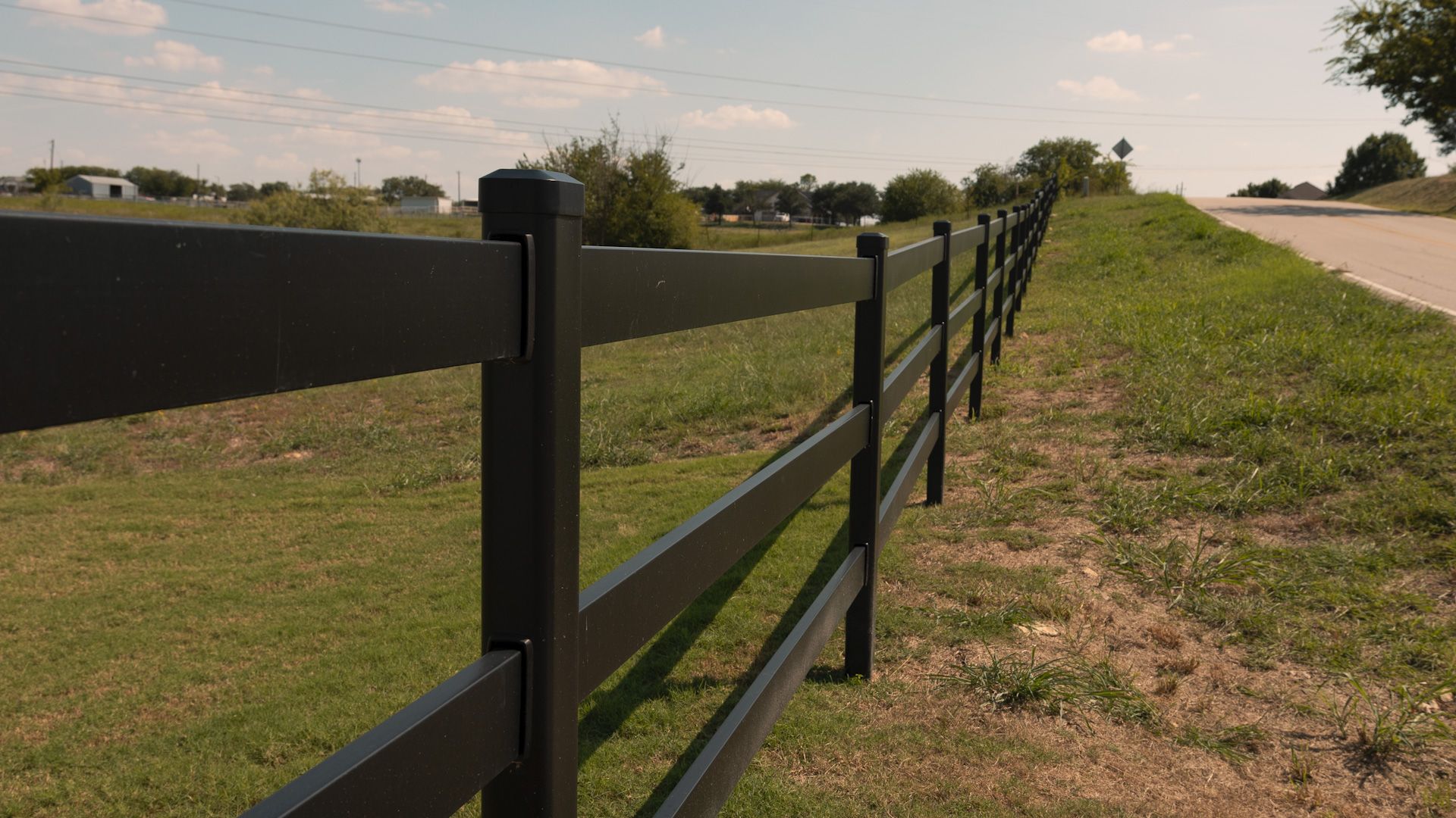 Black fence along a grassy area, with a road to the right, against a sunny sky.