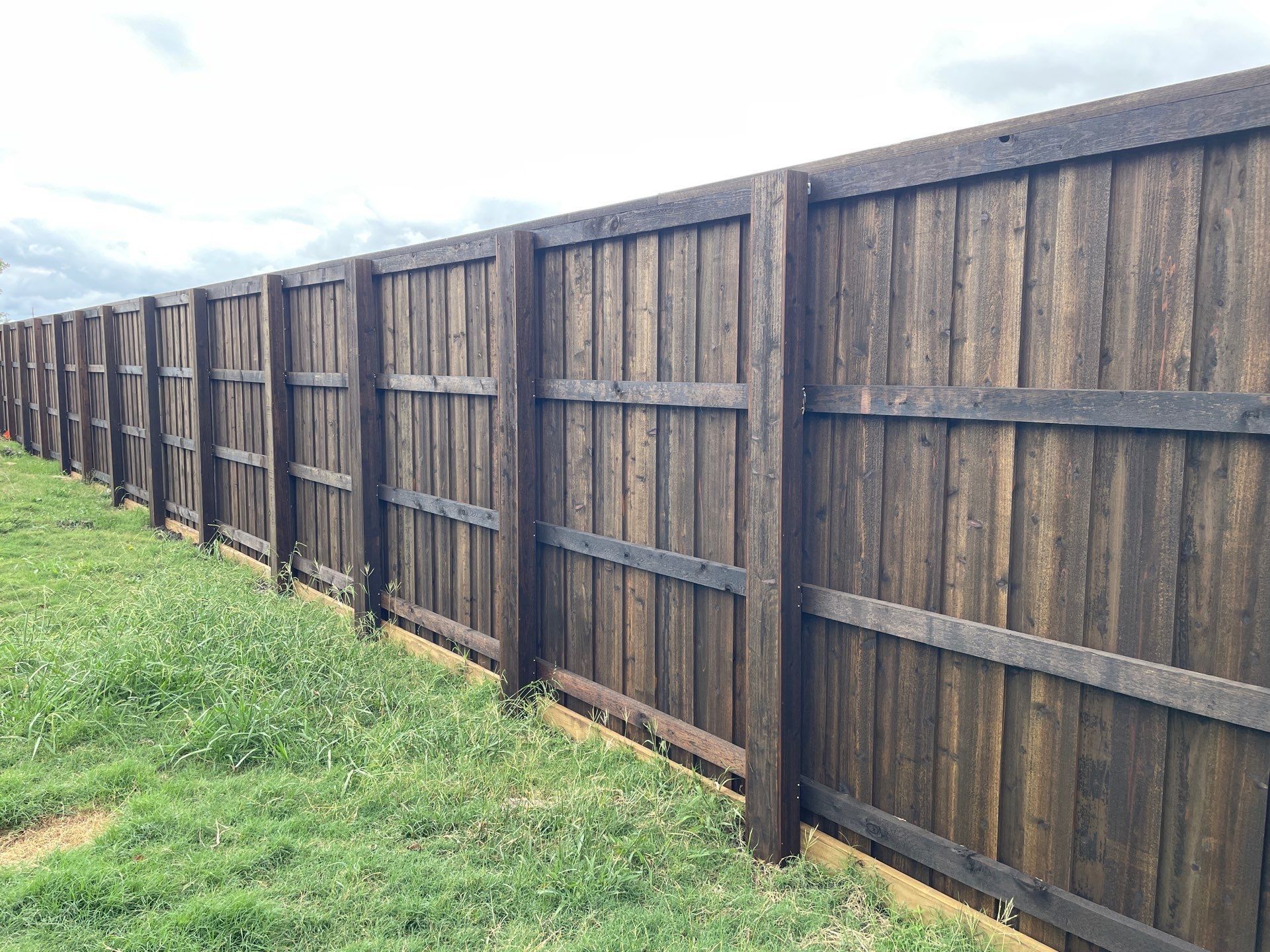 Wooden fence, stained dark brown, bordering green grass under a cloudy sky.