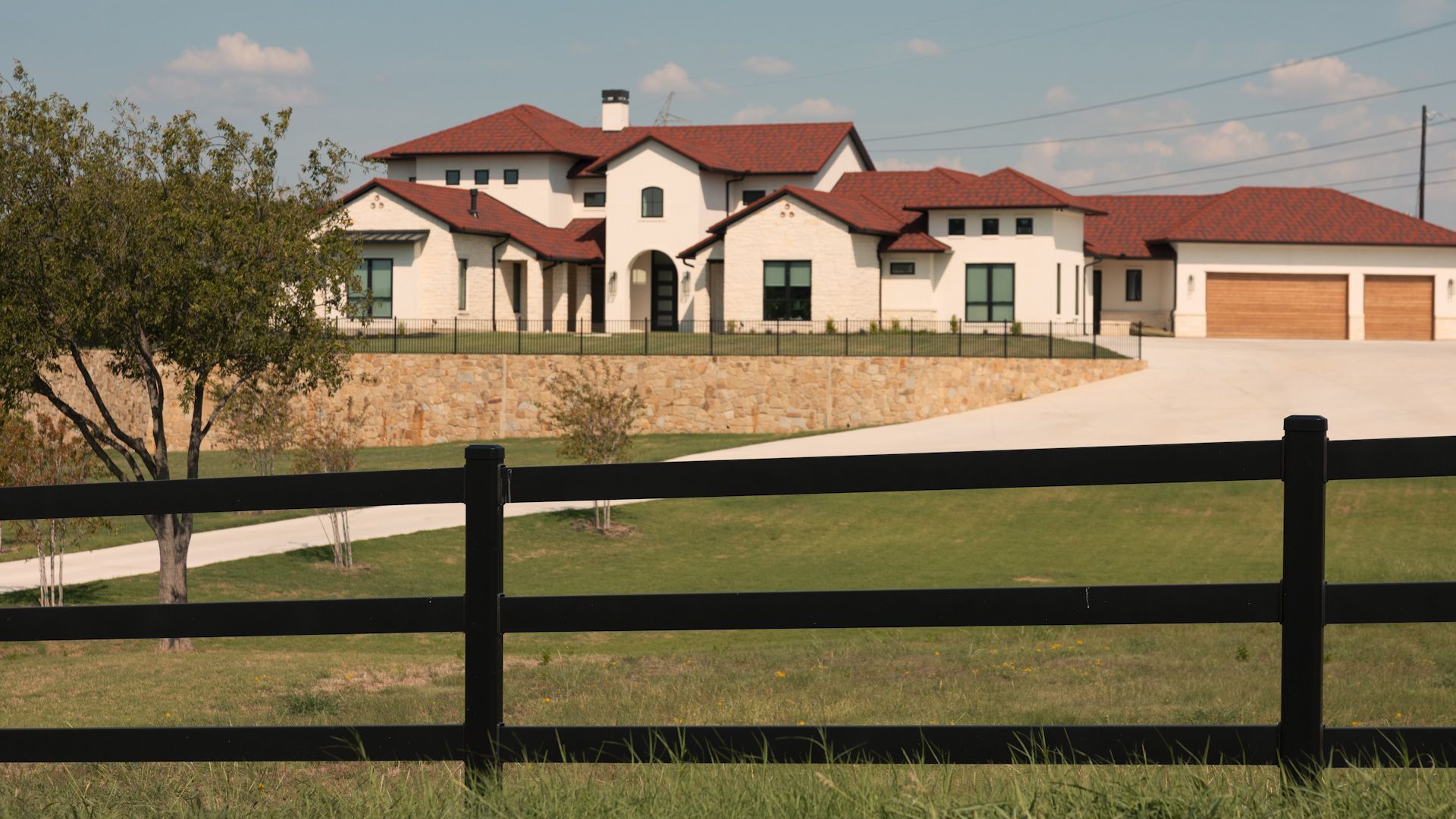 Large white house with brown roof, two car garage, set on a hill behind a black fence, sunny day.