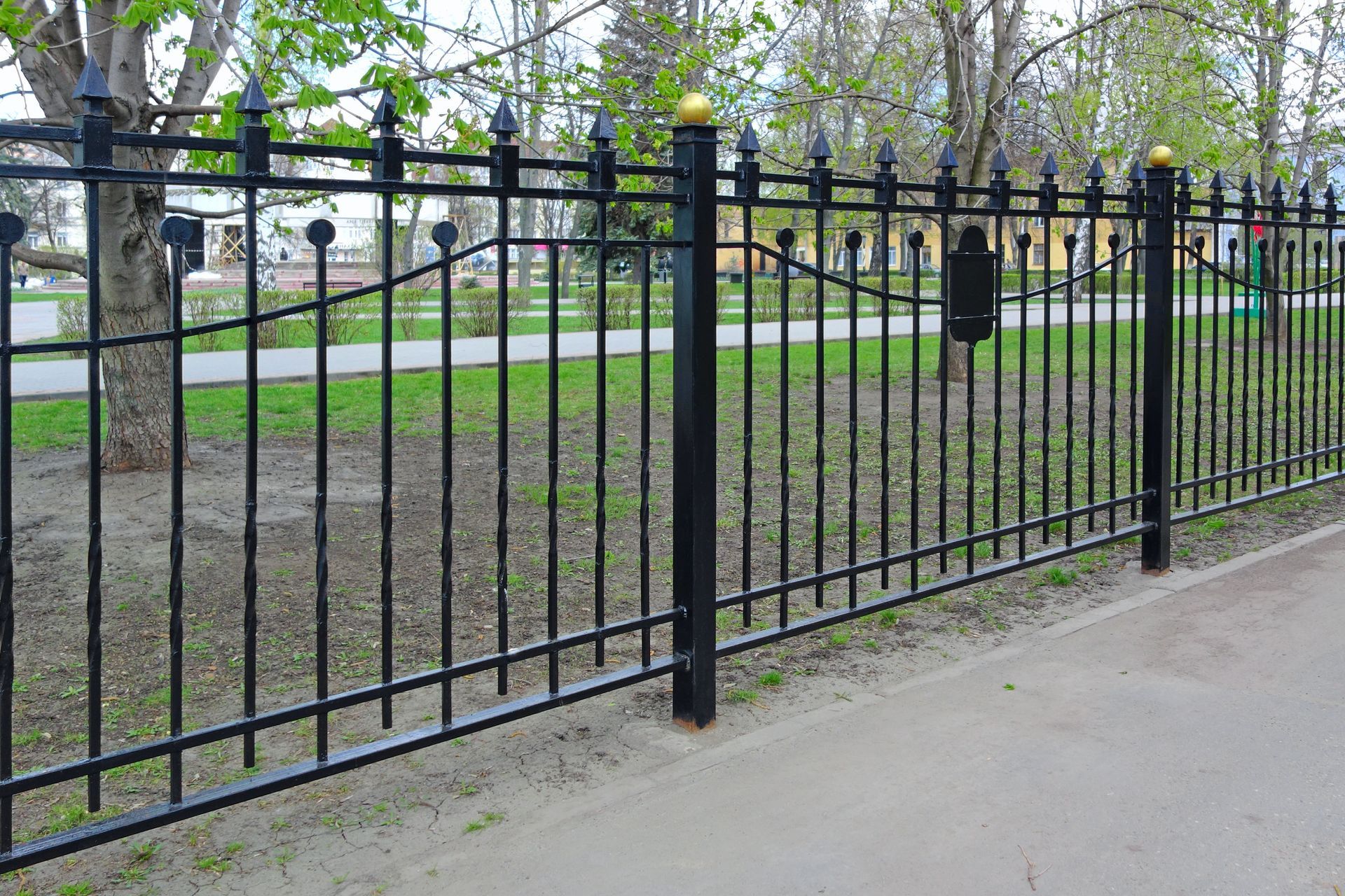 Black wrought iron fence along a sidewalk, with decorative finials and a grassy area in the background.