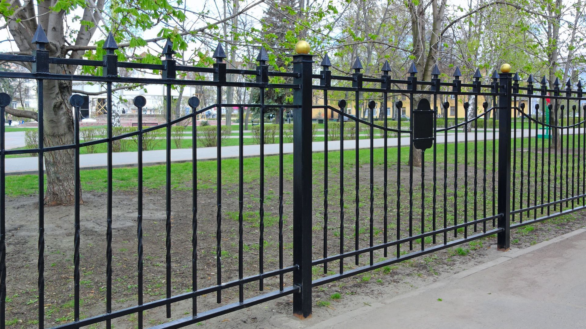 Black wrought iron fence bordering a grassy area with trees in the background.