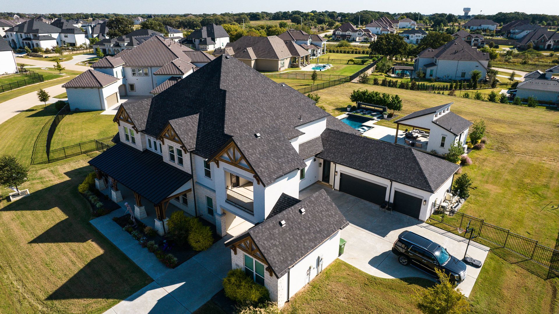 Large white house with dark gray roof, a black vehicle, and other houses in the background on a sunny day.