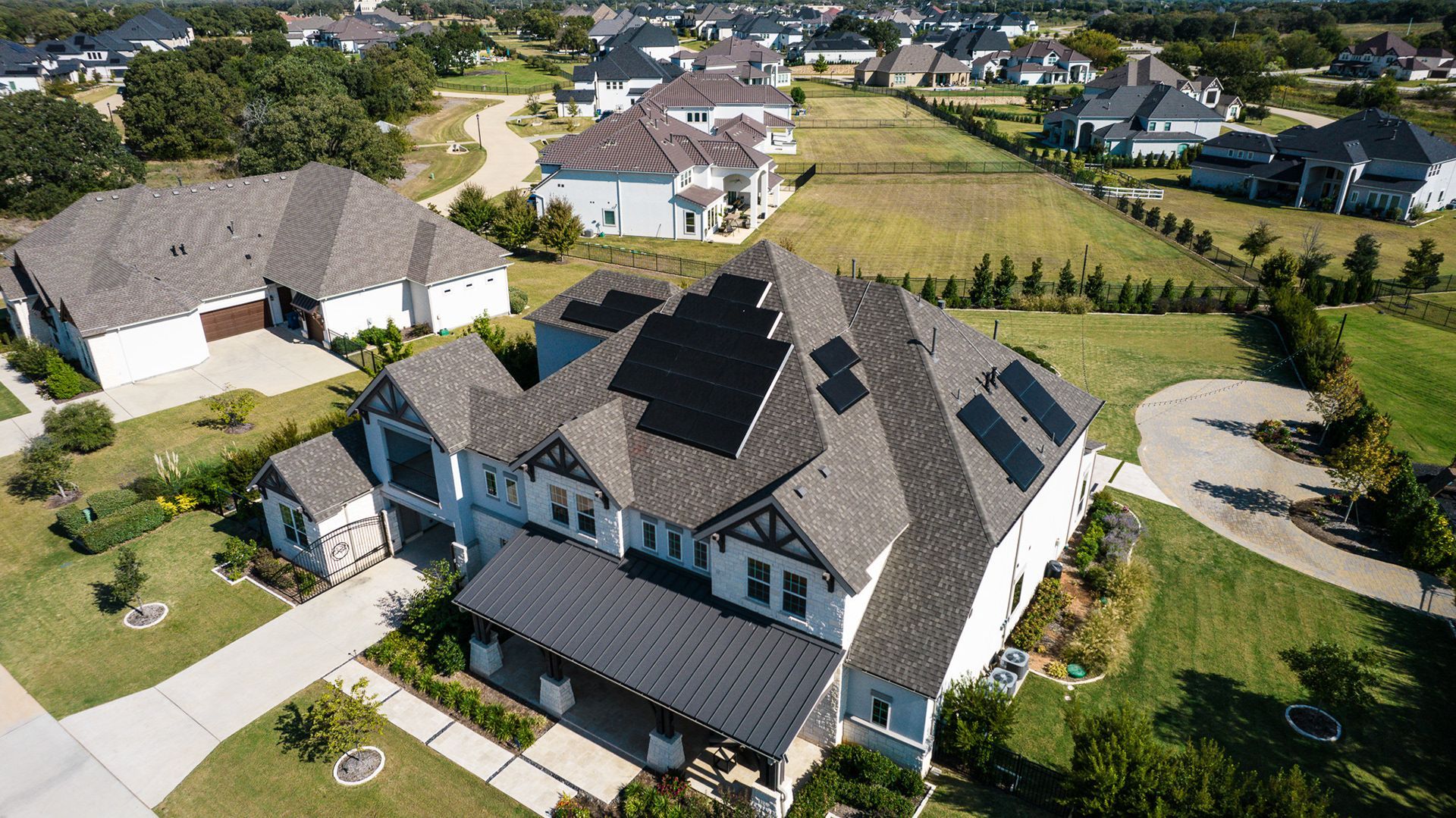 Aerial view of large white house with dark roof, front porch, and driveway in a suburban neighborhood.