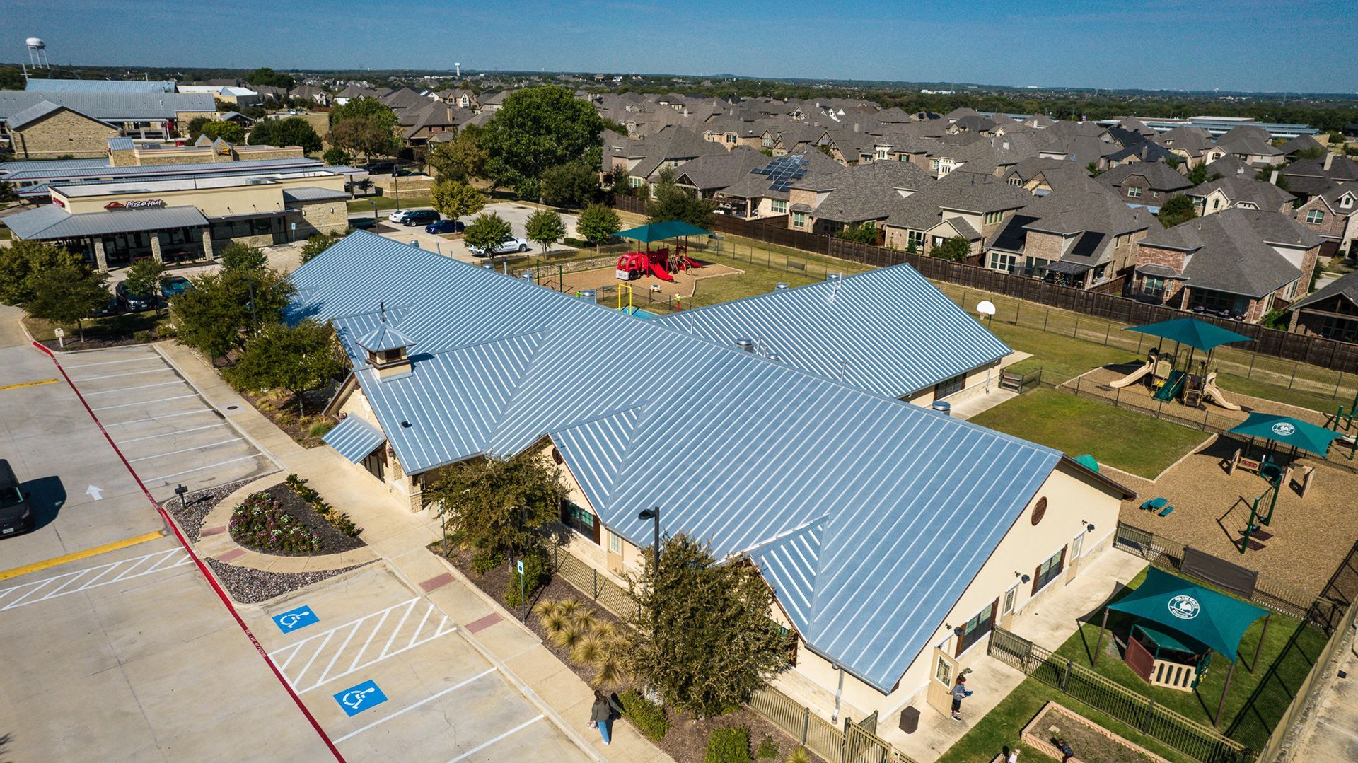 Aerial view of a light-colored, uniquely-roofed building with a playground and residential neighborhood in the background.