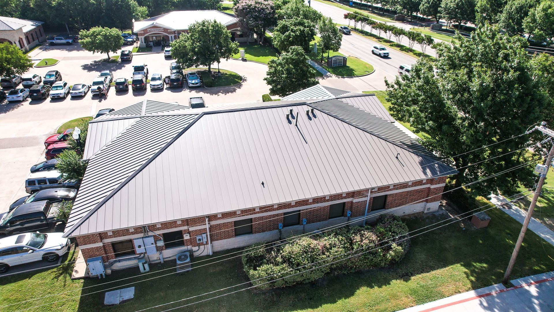 Aerial view of a brick building with a metal roof, cars parked in the lot, and trees around it.