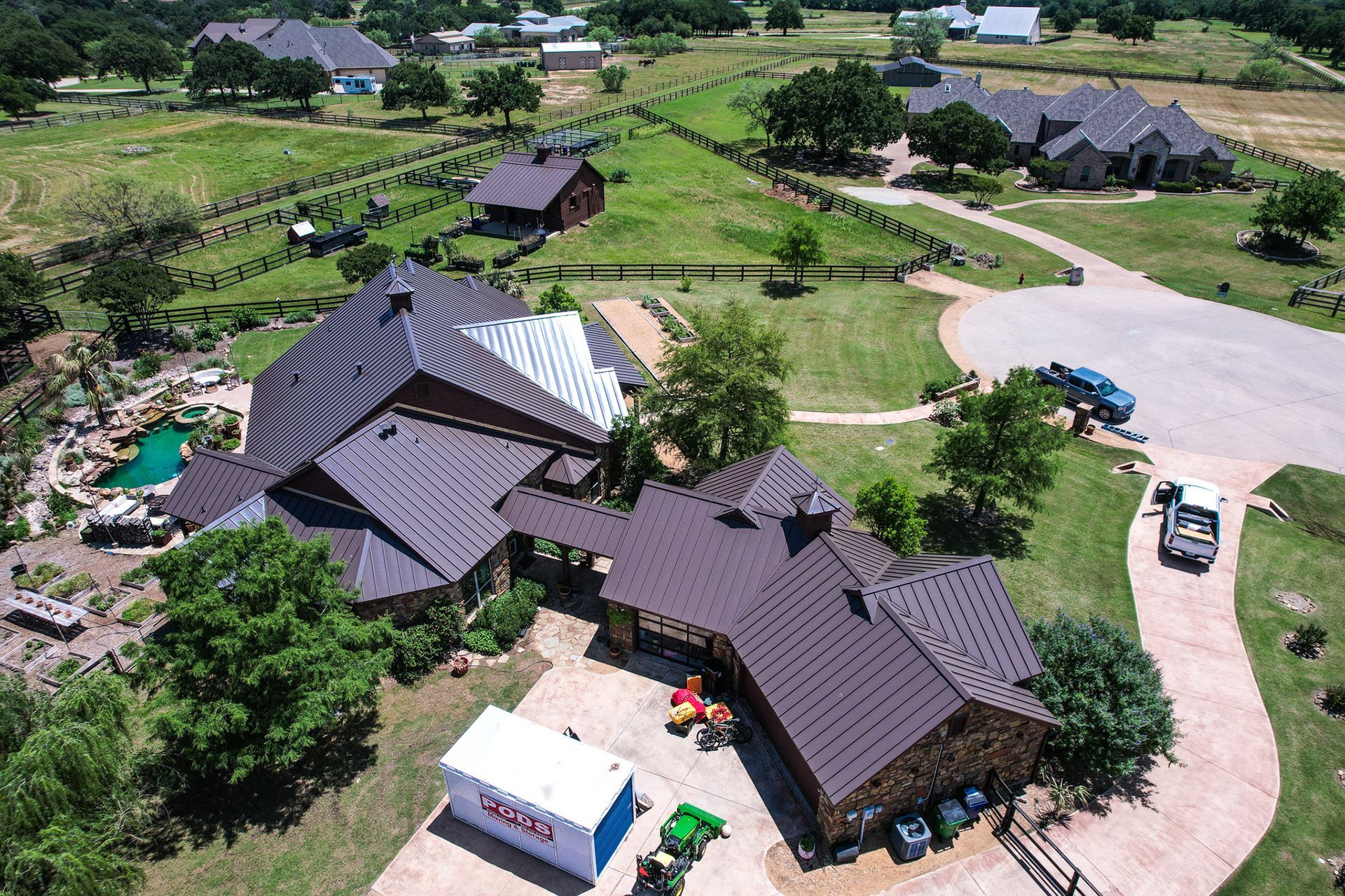 Aerial view of a large house with a unique roof design, surrounded by green fields and a pond.
