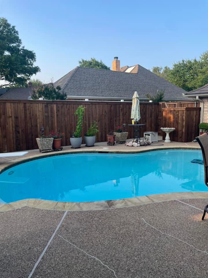 Pool in a backyard with a brown fence, potted plants, a table, and a birdbath; the sky is blue.