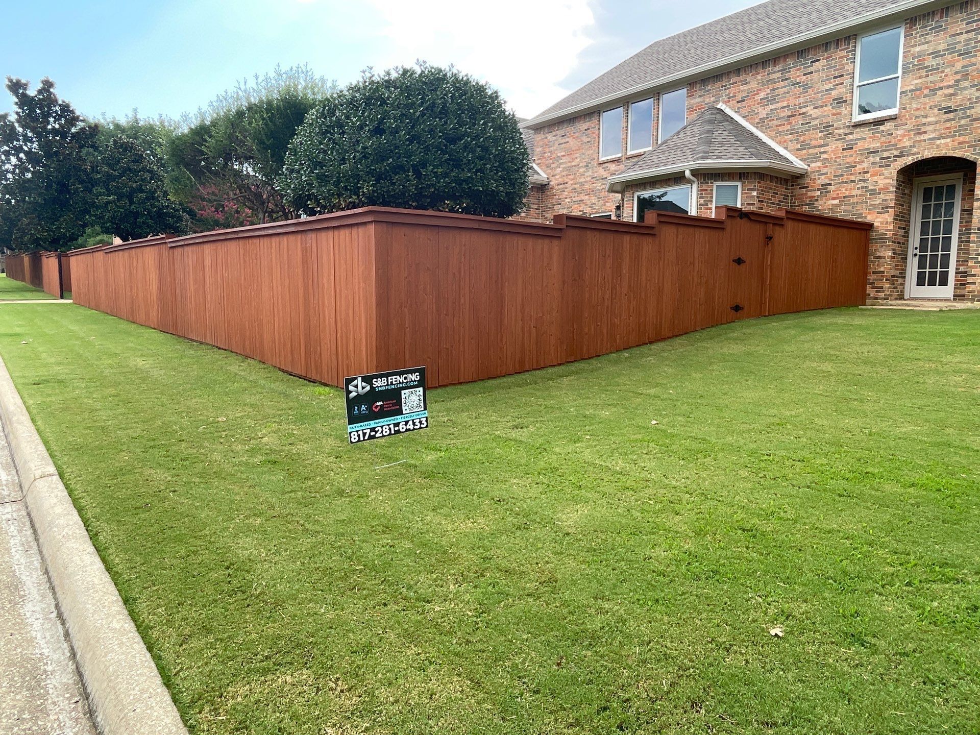 A brown stained wooden fence surrounds a lawn in front of a brick house.