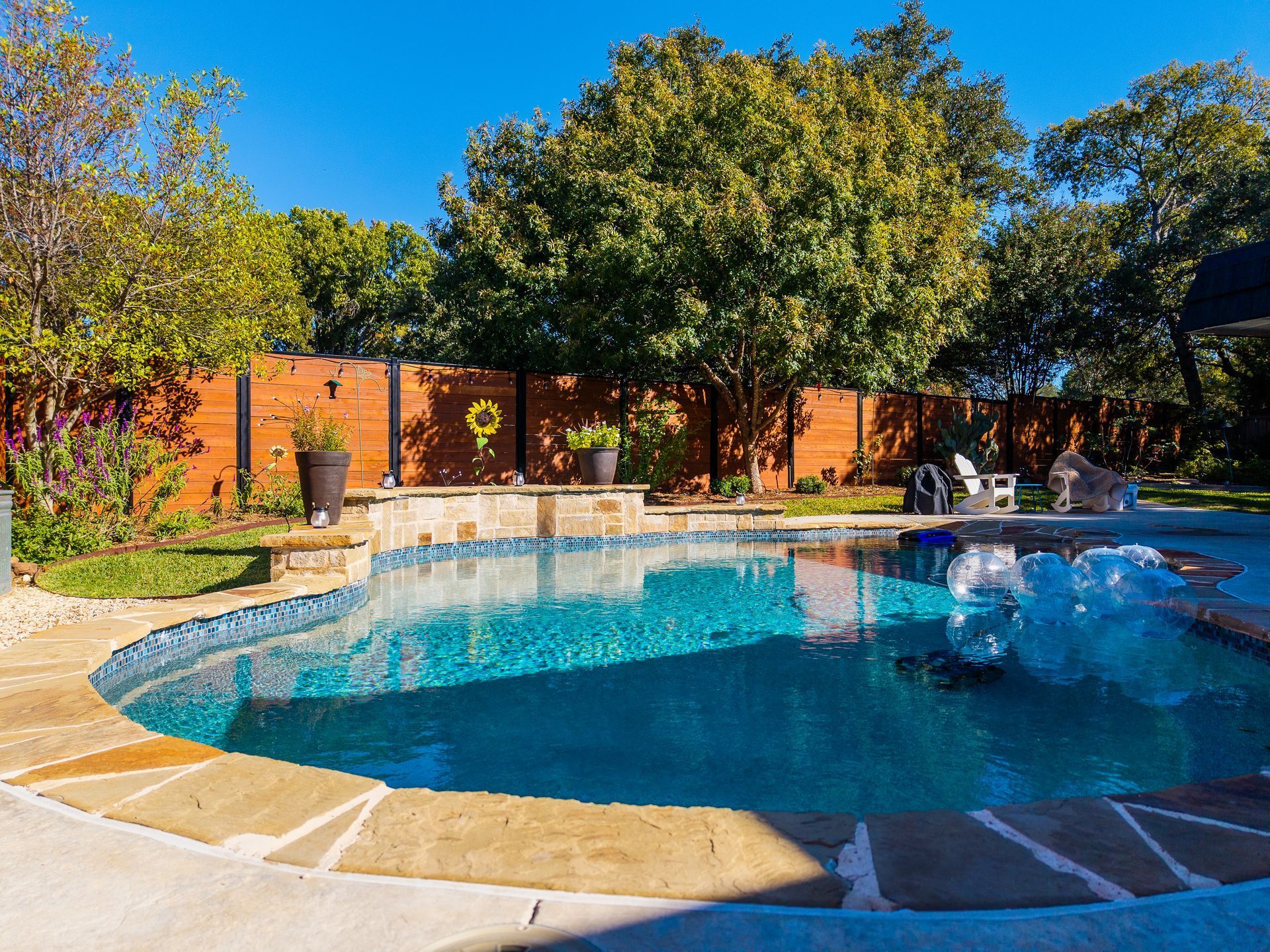 Pool with turquoise water, stone border, and wooden fence. Lush green trees and blue sky background.