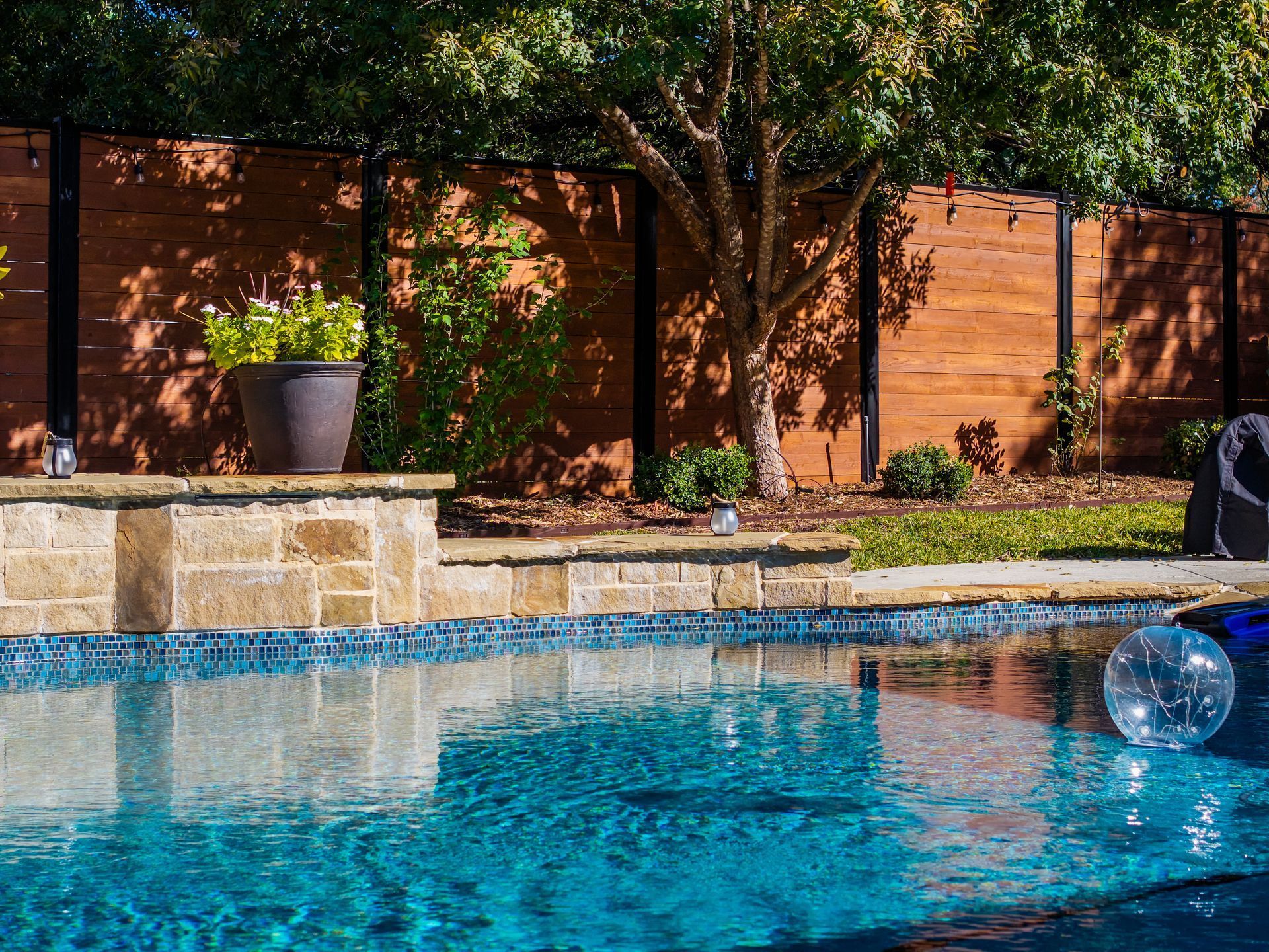 Swimming pool with blue tiles, stone border, and wooden fence in a backyard.