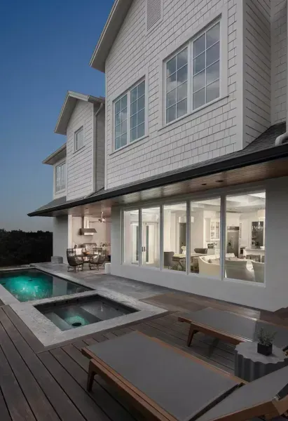 Exterior of a white house with a pool and hot tub. Wooden deck, glass windows, and lounge chairs.