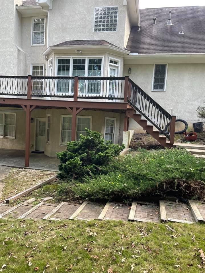 Back of a beige house with a wooden deck and staircase. Green bushes and a path lead from the yard to the house.