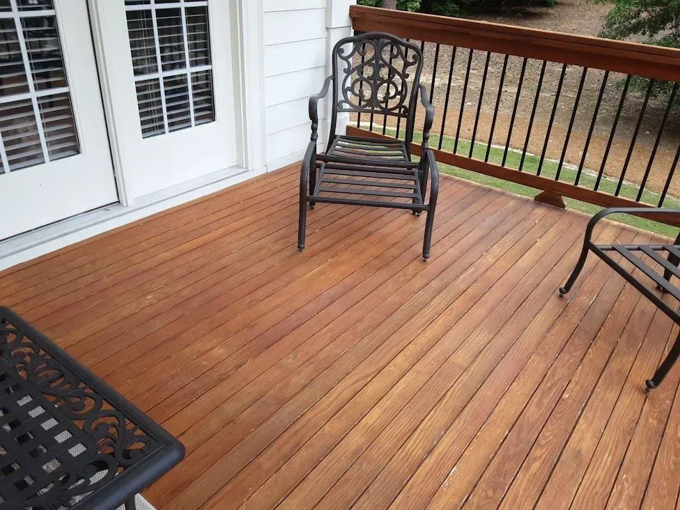 Wooden deck with two wrought iron chairs and a small table. Double white doors and a dark railing are visible.