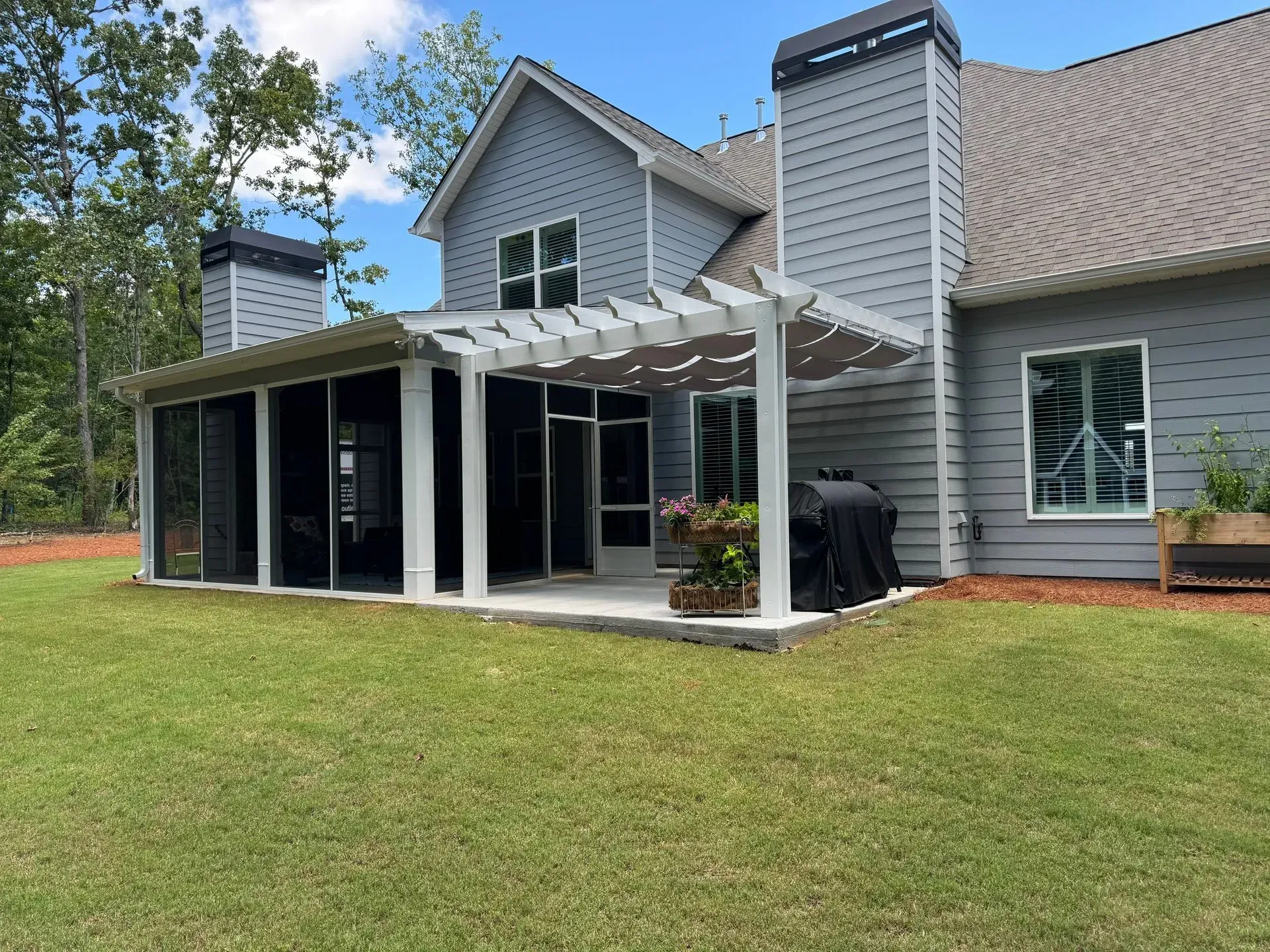 Grey house with a screened-in porch and pergola, surrounded by green grass and trees, under a blue sky.