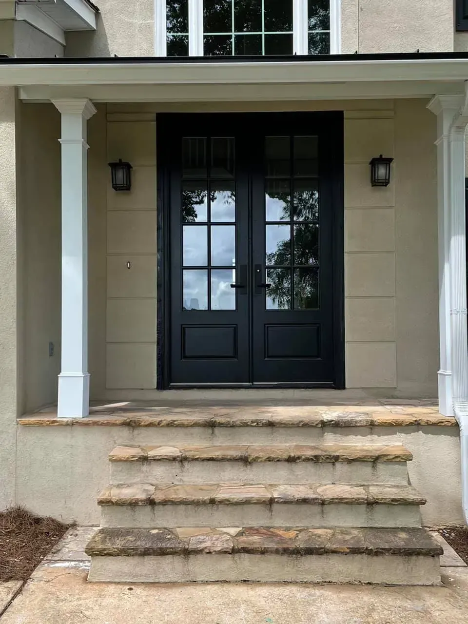 Black double doors with glass panes, flanked by white columns and sconces, leading to a home with stone steps.