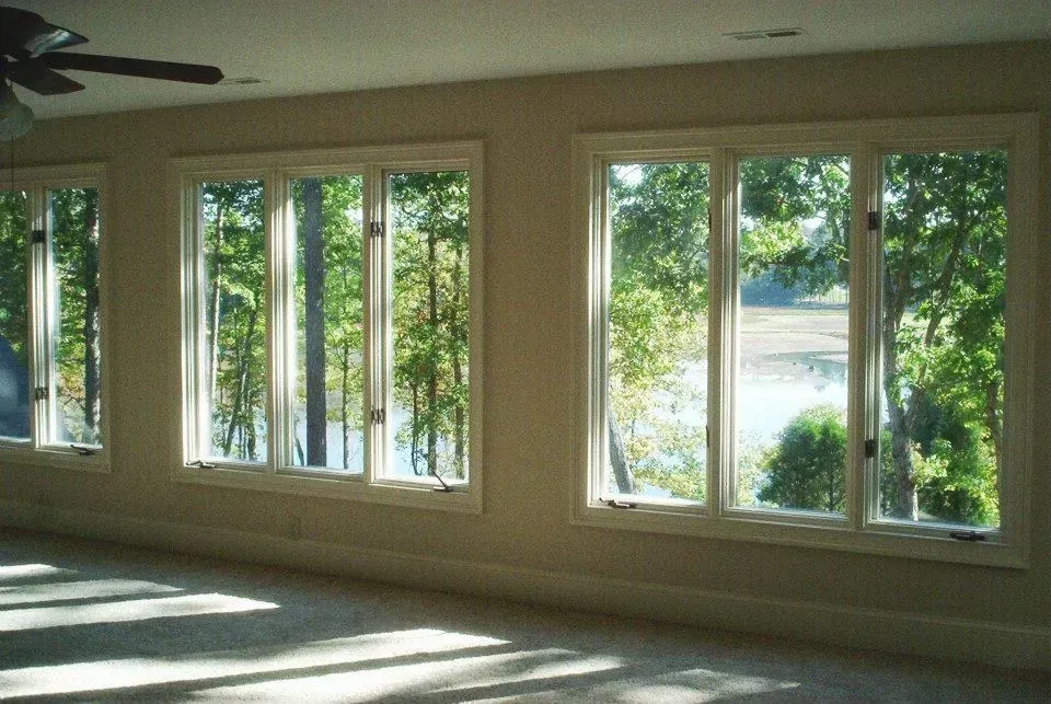 Three white-framed windows overlooking a lake, with trees in the background. Sunlight streams into the room.
