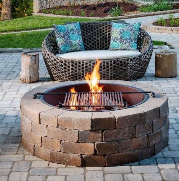 Fire pit with grill, wicker loveseat with pillows, and wood stools on a stone patio.