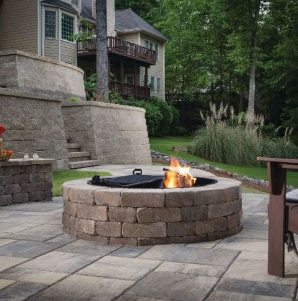 Brick fire pit with flames, on a stone patio, in front of a multi-level house with landscaping.