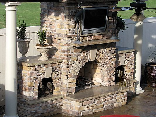 Outdoor stone fireplace with TV, archway, and wood storage. Beige and tan hues on a patio setting.
