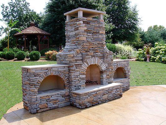 Stone outdoor fireplace with arched openings and chimney on a patio.