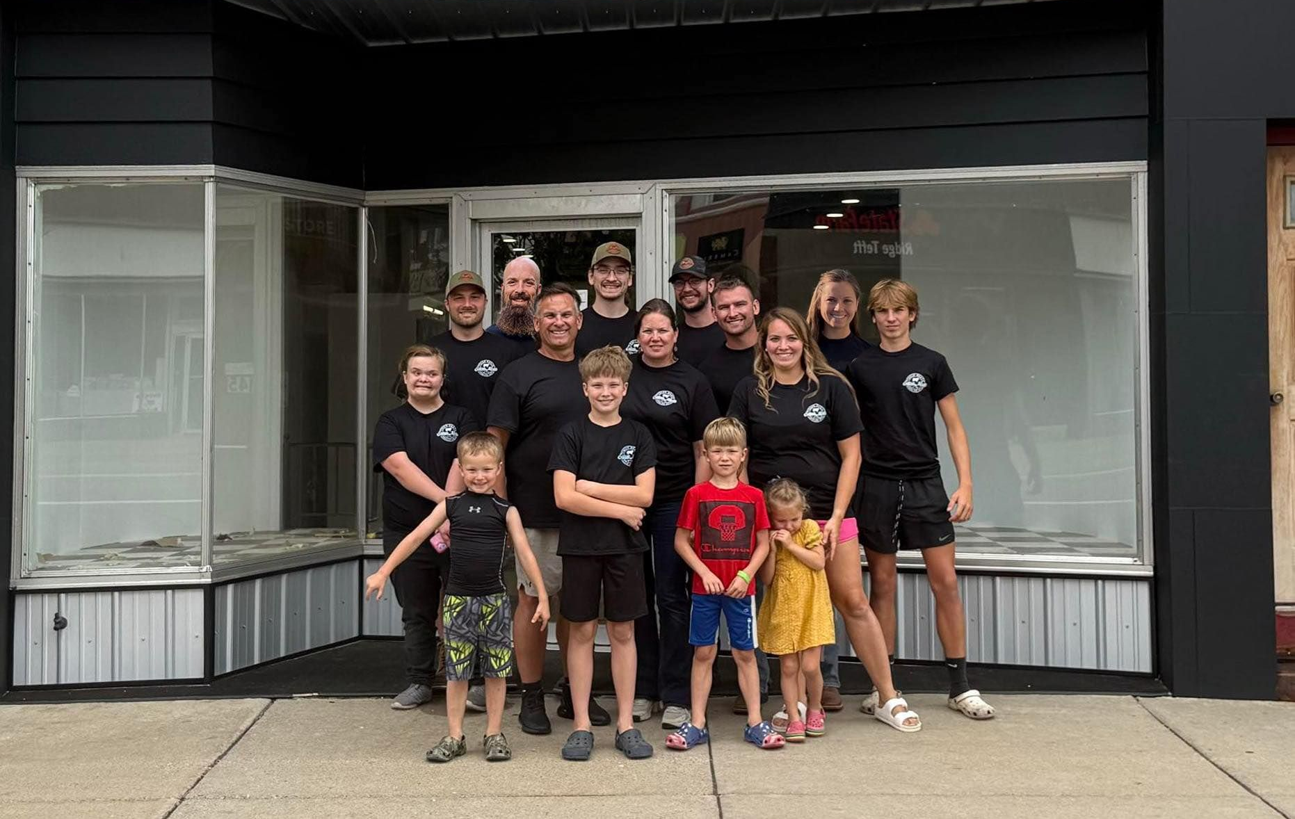People pose in front of "Eaton Meat" store sign. Black building, sunny day.