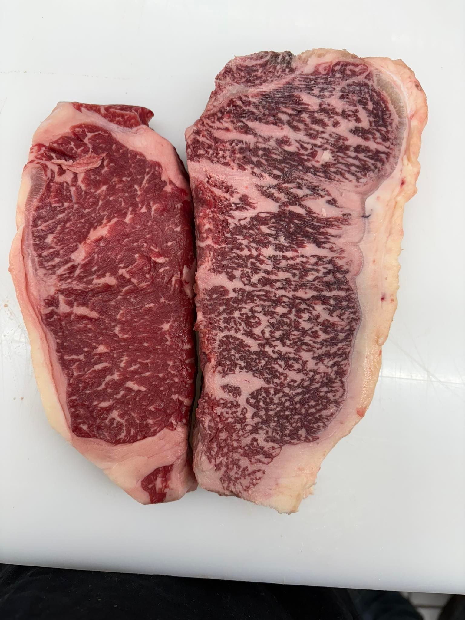 Two raw strip steaks on white surface, one with heavy marbling.