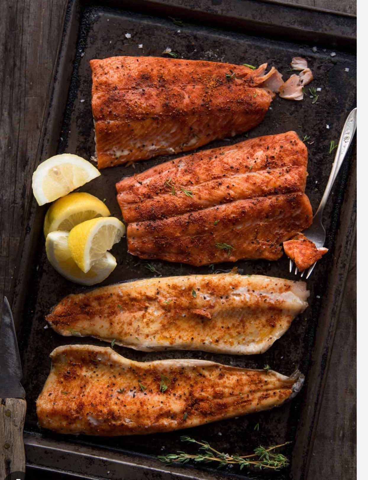 Grilled fish fillets on a dark metal tray with lemon wedges and a fork, seasoned.