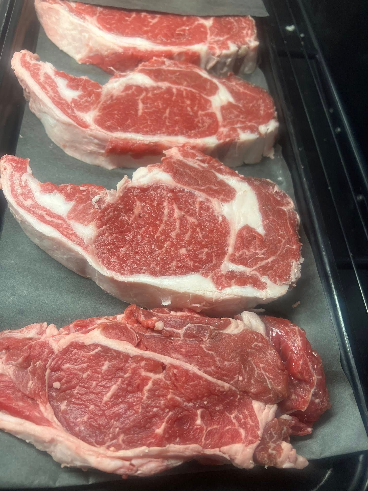 Four raw ribeye steaks on a black tray.