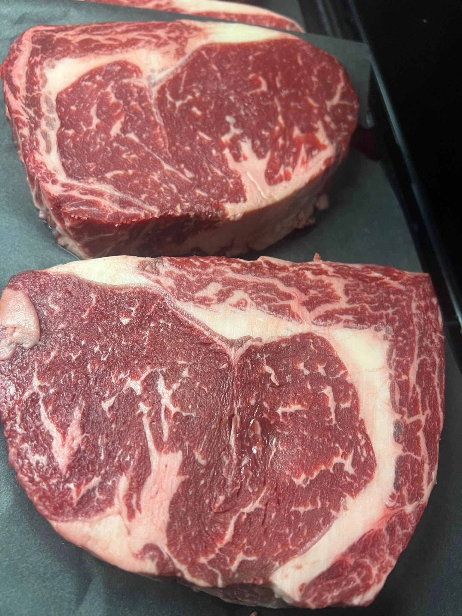 Two raw ribeye steaks, showing marbling, resting on a dark surface.