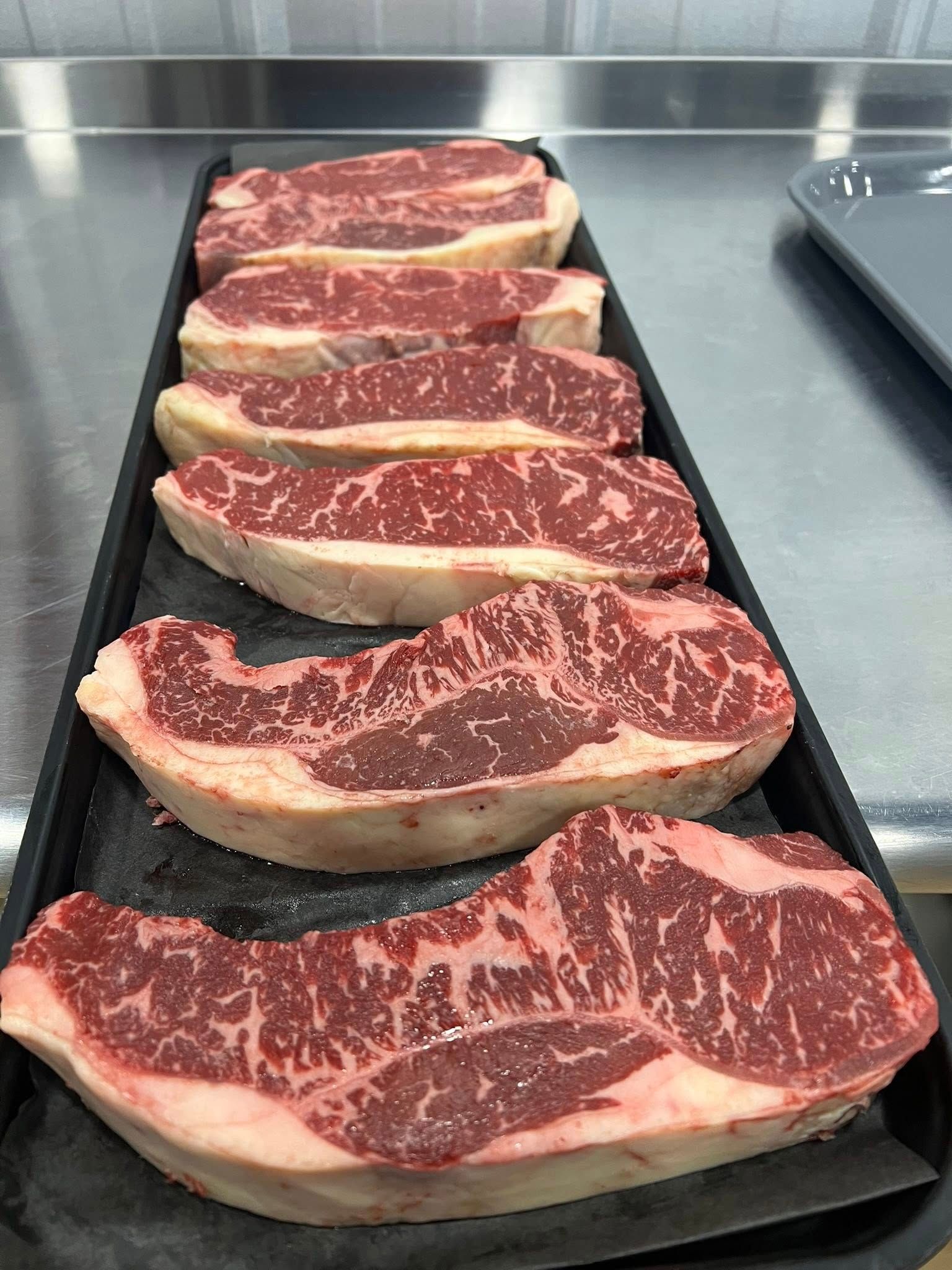 Raw steaks arranged in a black tray on a metal surface, showing marbling and fat.