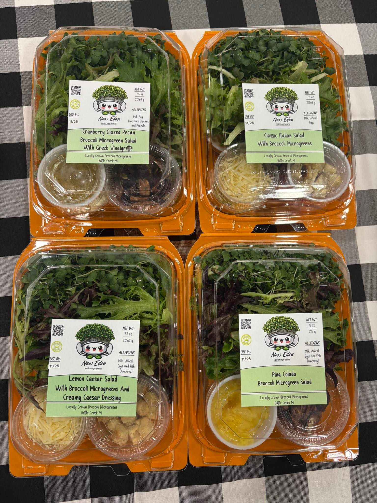 Four salad containers with greens, dressings, and toppings on a checkered tablecloth.