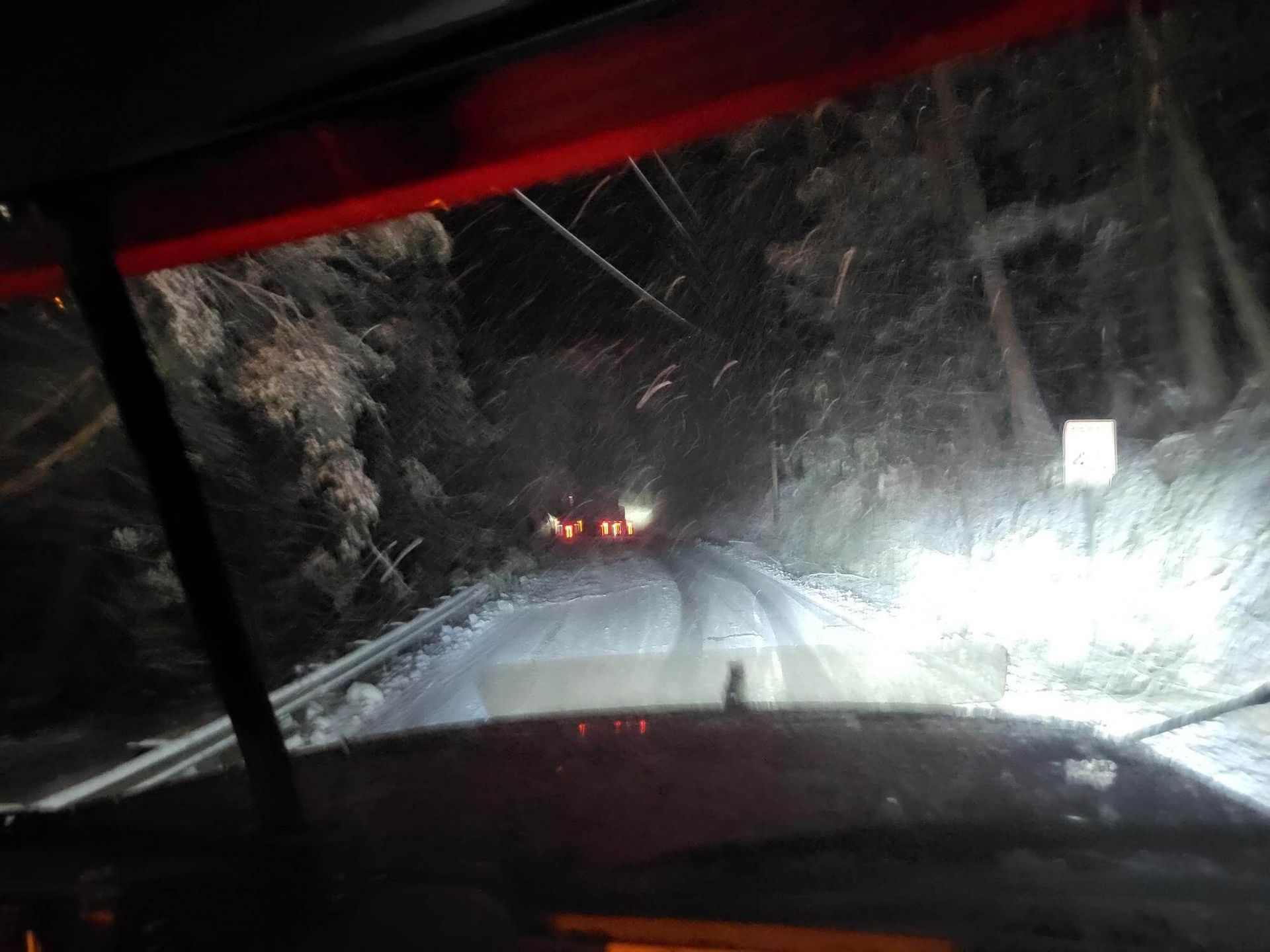 Snowy nighttime road seen from a car, headlights illuminating a narrow forest lane with distant taillights.