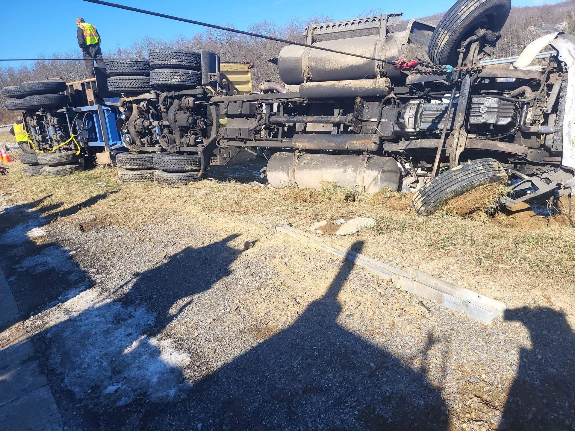 Overturned cement mixer truck on its side beside a road, with debris and two workers nearby.