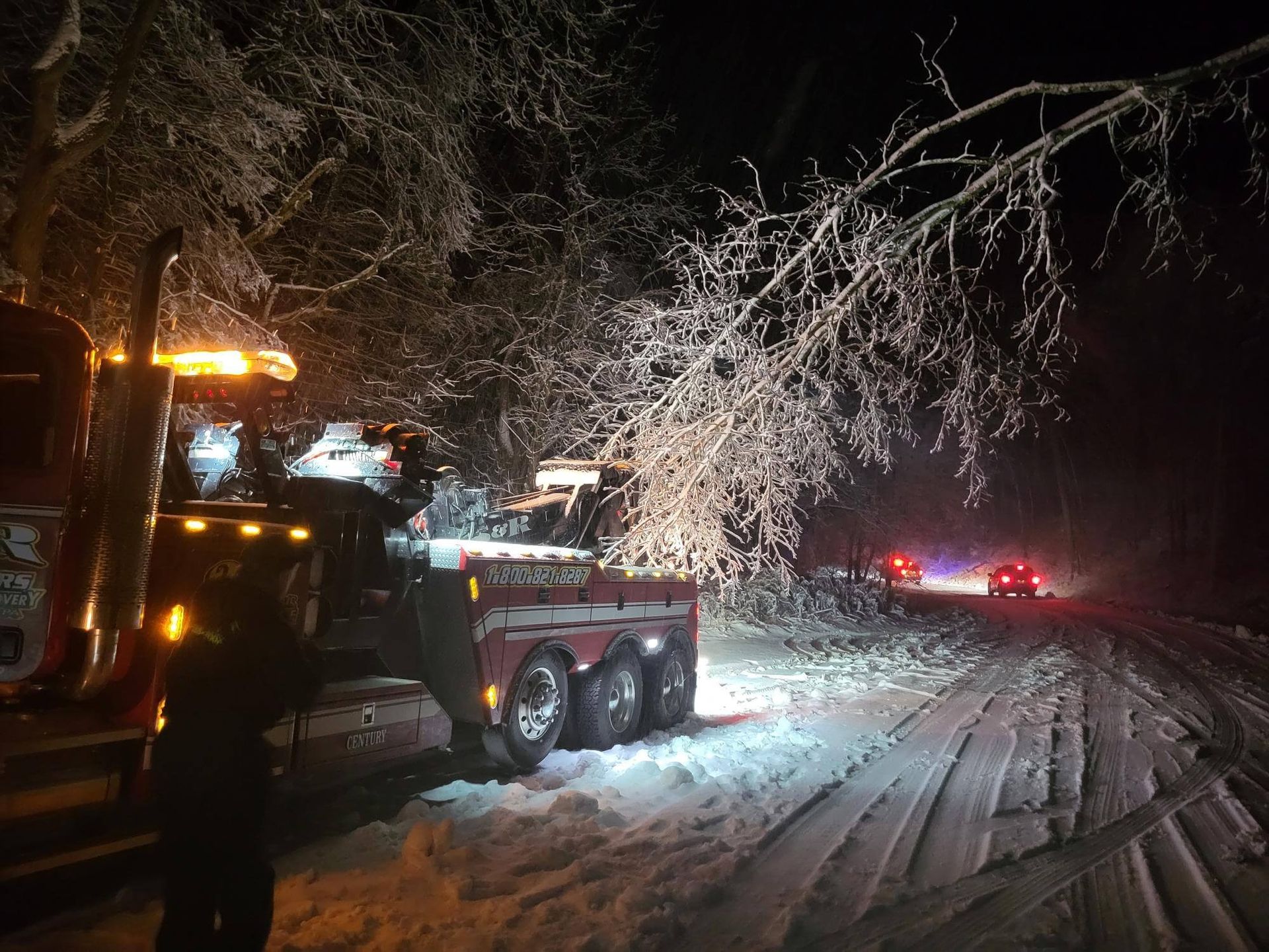 Snowplow clearing a snowy road at night, with a truck and red taillights ahead.