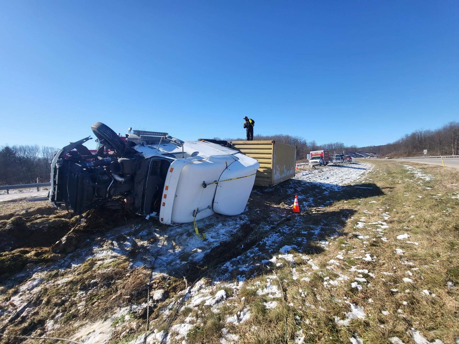 Overturned truck trailer on snowy roadside with debris and a person standing nearby under a blue sky
