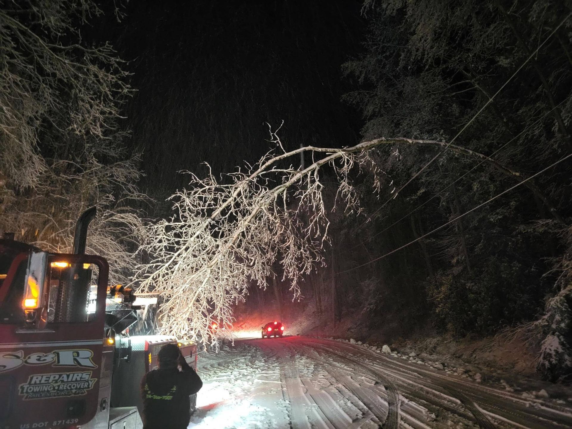 Snowy road blocked by fallen tree branches at night, with a tow truck and workers clearing the scene